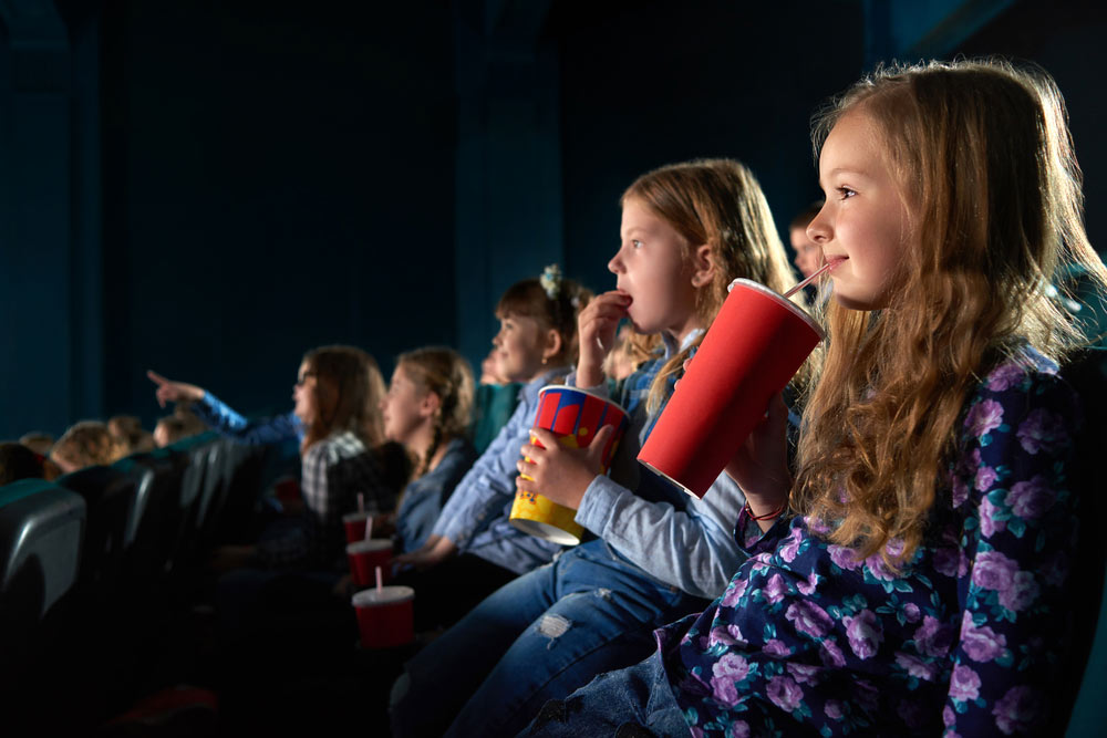 Cute young girl smiling joyfully sipping her drink watching a movie at the cinema