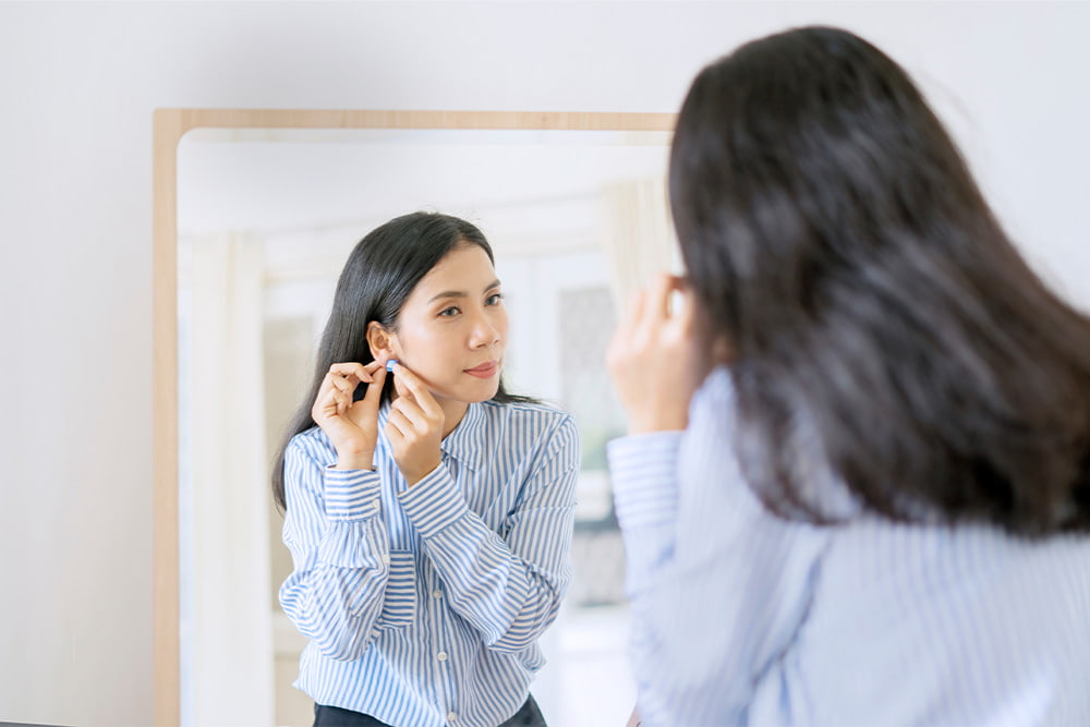 photo of business woman in blue striped button up shirt putting in earring in front of mirror while getting ready for work
