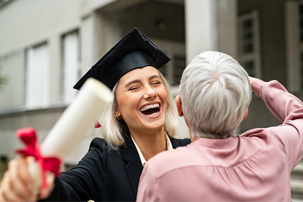 Happy female graduate in black cap and gown hugging grandmother while holding diploma and smiling