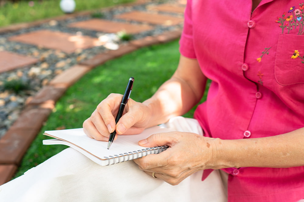 older woman writes in notebook with pen in grass