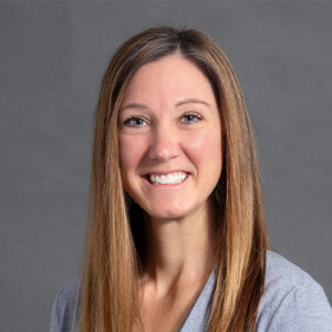 Smiling brunette with straight hair wearing a blue shirt against gray backdrop