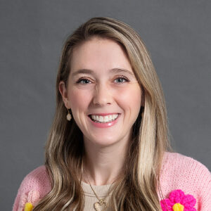 Woman with long blonde hair wearing a pink sweater with flowers smiling in headshot