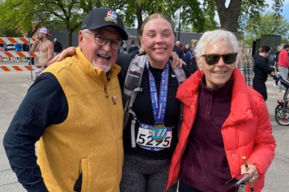Nancy and her husband on either side of their granddaughter wearing a medal and race bibs at the finish line of the Lincoln Half Marathon
