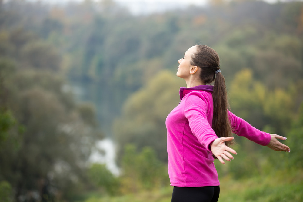 Woman standing outdoors while stretching her arms, enjoying nature in a pink jacket with a blurred forest background.