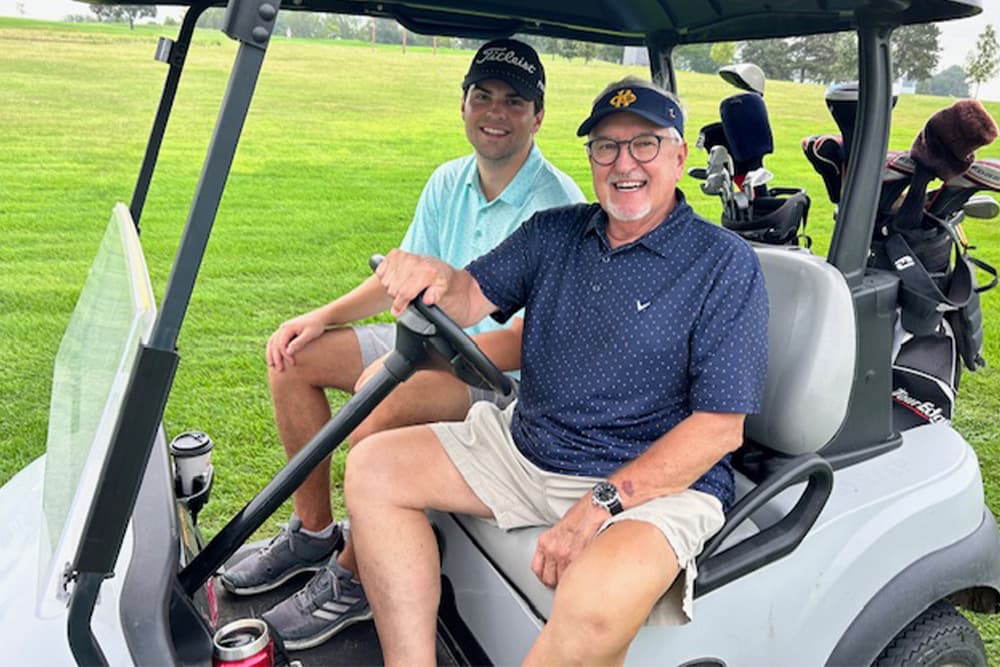 A man and his grandson smiling and sitting in a golf cart on a green golf course. The man in the driver's seat is wearing a navy blue shirt, glasses, and a dark baseball cap, while the younger man beside him is wearing a light blue shirt and a dark hat. Golf bags are visible in the back of the cart.