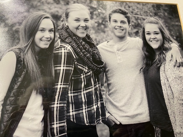 Black and white photo of Nancy's grandkids posing in fall, with three granddaughters and one grandson with arms wrapped around each other's shoulders