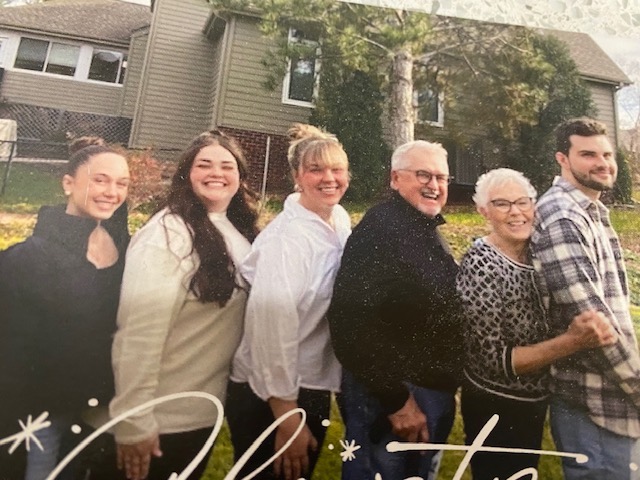 Nancy and her husband John pose in a line with her three granddaughters and one grandson, all smiling, in the backyard for Christmas