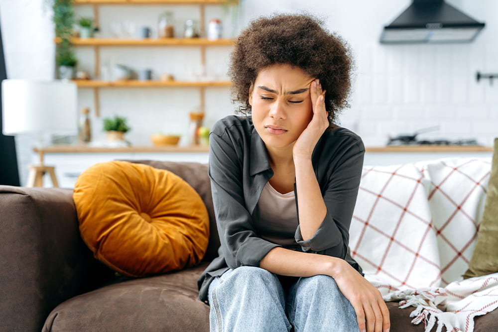 Frustrated, young African American woman in casual clothes, sitting on the sofa in the living room, pressing her hand to her temple