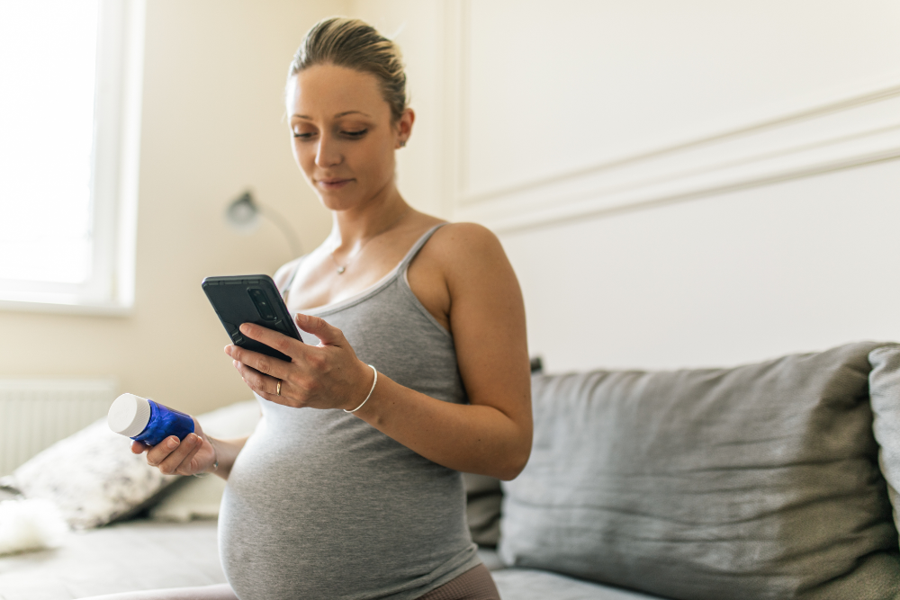 pregnant woman sitting on bed, holding her phone and a bottle of medication